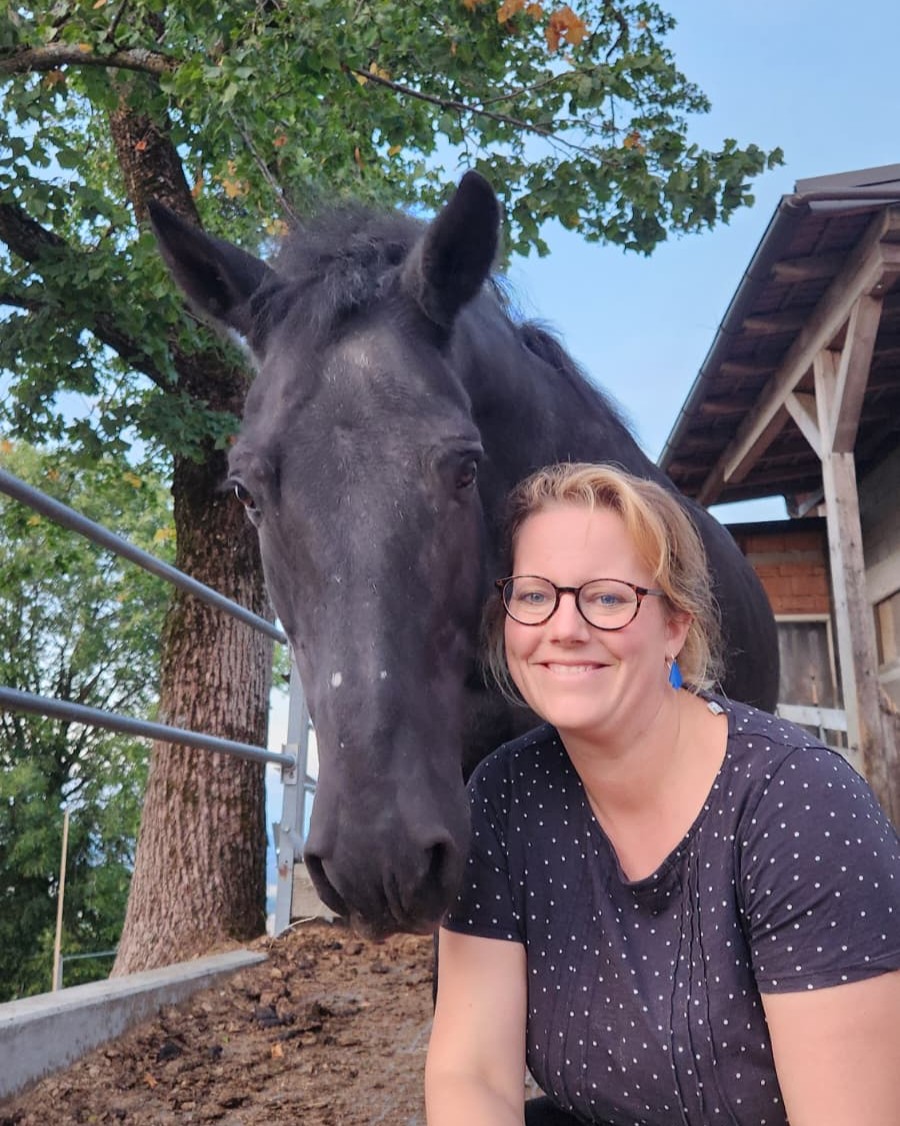Birgit lächelt eng an ihr schwarzes Pony Merlin gelehnt, im Hintergrund Bäume und der Stall des Erlebnishofs